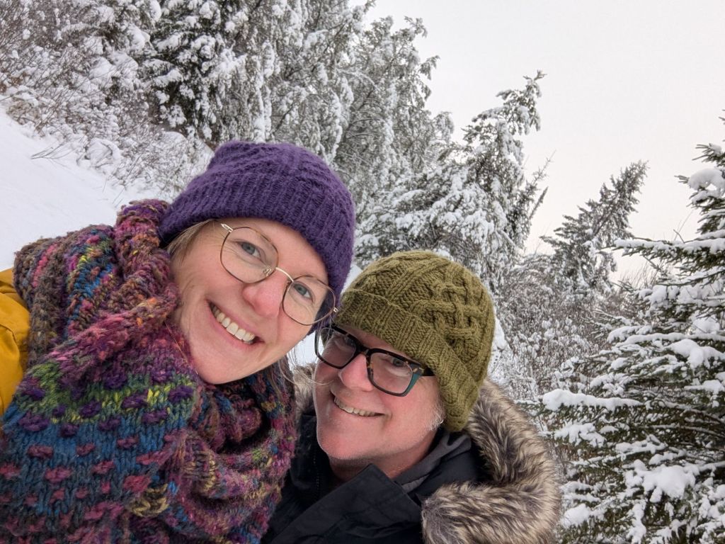 A selfie of Amy and Jake, smiling and bundled up in parkas and knitted winter toques made by Amy. Amy is also wearing a colourful handknitted scarf she made. Amy wears her thin, tortoise-shell and gold rimmed glasses, and Jake wears his thick tortoise shell and green rimmed glasses. They are surrounded by snow-covered pine trees at Falcon Trails Resort. The sky is overcast and white. They are white Canadian settler-decendants in their early 40s and 50s. Photo taken December 2024.